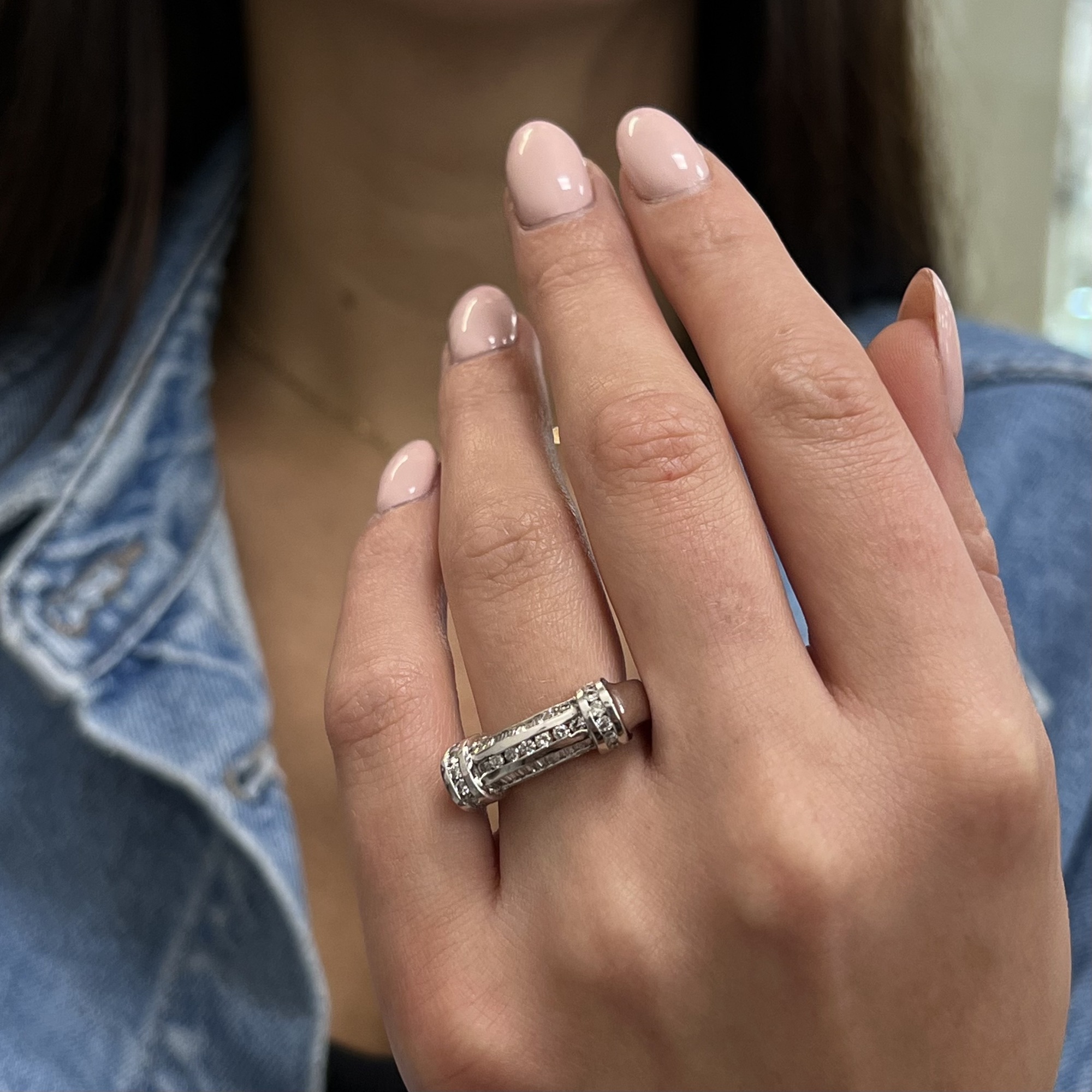 An image of a Rachel Koen women's ring with diamonds, worn on the ring finger of a person's left hand. The hand is positioned with a slight tilt towards the camera, allowing a clear view of the ring's top and side. The ring features a detailed band design with multiple sparkling diamonds set in a white gold. The close-up shot captures the ring from a front-facing angle at a moderate distance, emphasizing the ring's intricate details and the reflective shine of the diamonds.The ring is pre owned in a great condition.