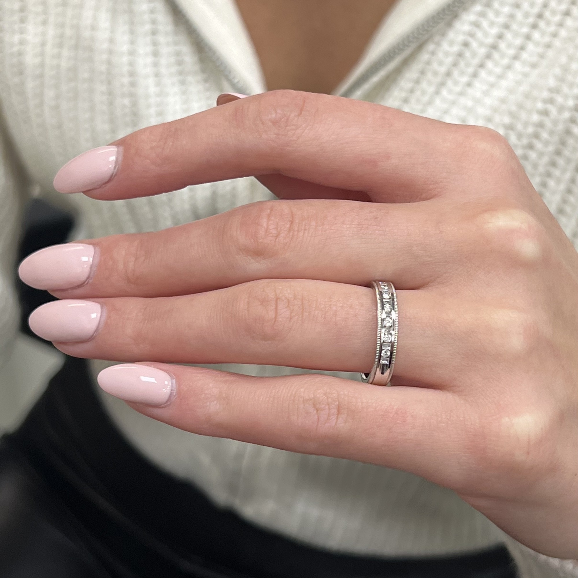 An image of a woman's hand presenting a Rachel Koen brand ring with diamonds. The ring is positioned on the ring finger, close-up at a slight angle, showcasing the band's design. The band is white gold, with a row of small, round-cut diamonds encircling it. The wearer has light pink polished nails, and the background is softly blurred.The ring is pre-owned in a great condition.The ring is pre-owned in a great condition. 