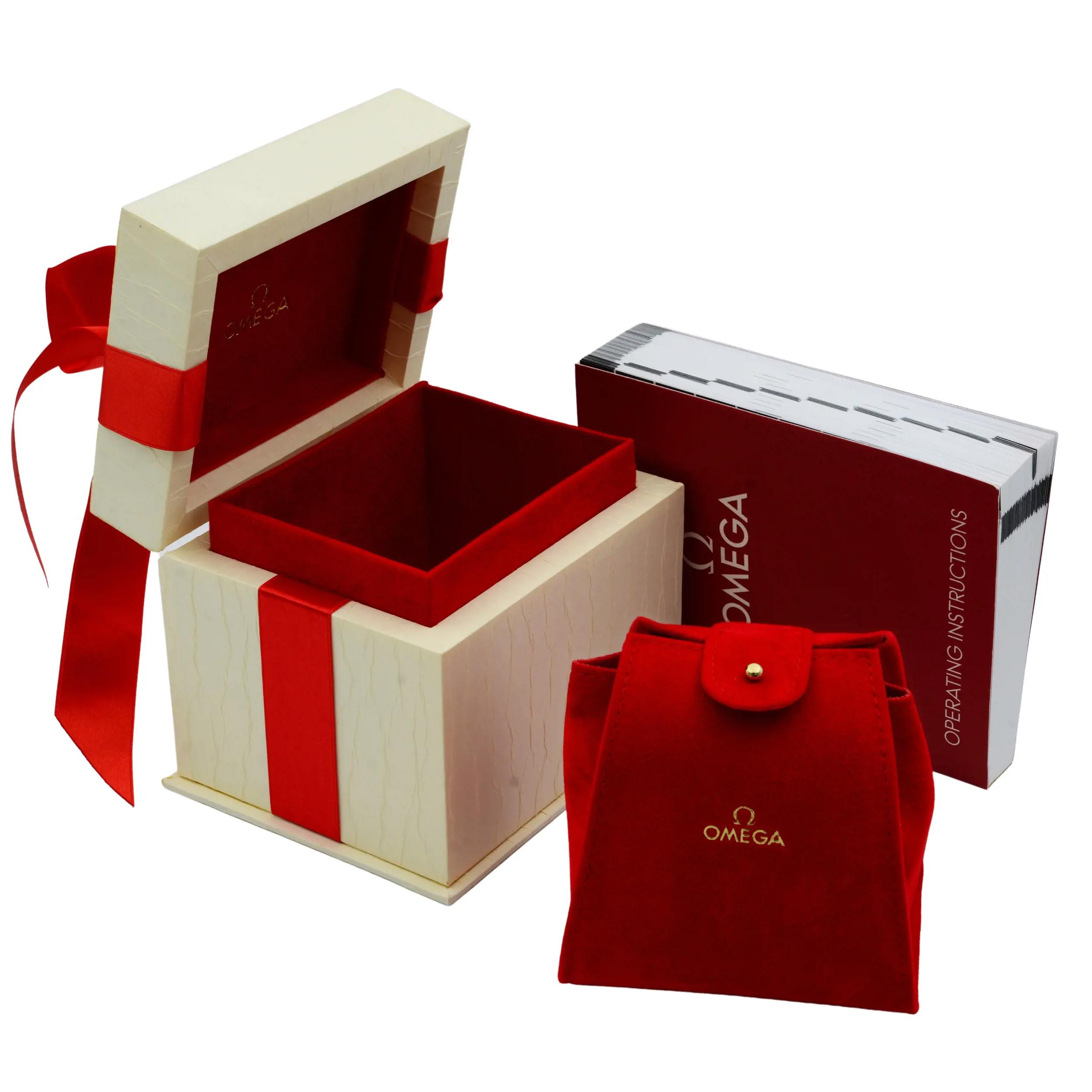 An image of a wristwatch display model packaging. The image shows an open wooden box with a cream-colored exterior and a red interior lining, positioned centrally. A red ribbon decorates the box. To the right, a red velvet pouch stands upright with a buttoned flap. Behind the pouch, a red booklet titled "Operating Instructions" is positioned upright. The angle is slightly elevated, capturing the items from a short distance, highlighting the luxurious presentation.
