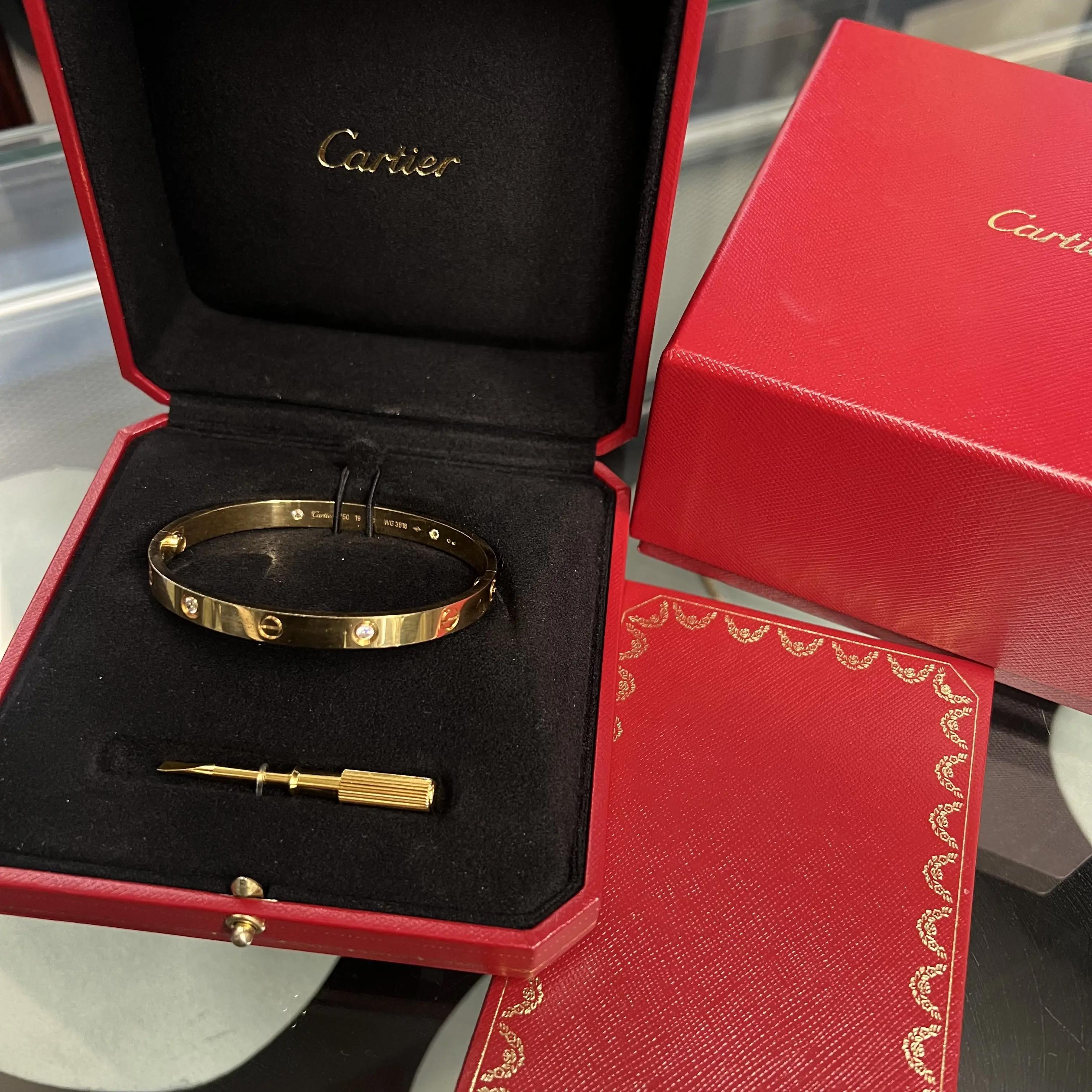 An image of a Cartier love unisex adult bracelet with diamonds, displayed at an oblique angle and positioned centrally in an open, velvet-lined presentation box, which is resting on a glass surface. A yellow gold bangle with evenly spaced diamond accents is in the foreground, complemented by a cylindrical screwdriver lying below it within the box. The iconic Cartier logo is embossed in gold on the inner lid of the box, which is slightly tilted towards the viewer, enhancing the visibility of the bracelet. To the right, a partially visible matching red Cartier box with gold trim sits beside the open box, providing a sense of the brand's signature packaging. The image is taken from a moderate distance, allowing for the full display of the open box and the bracelet's exquisite details to be appreciated. Comes in a great preowned condition.