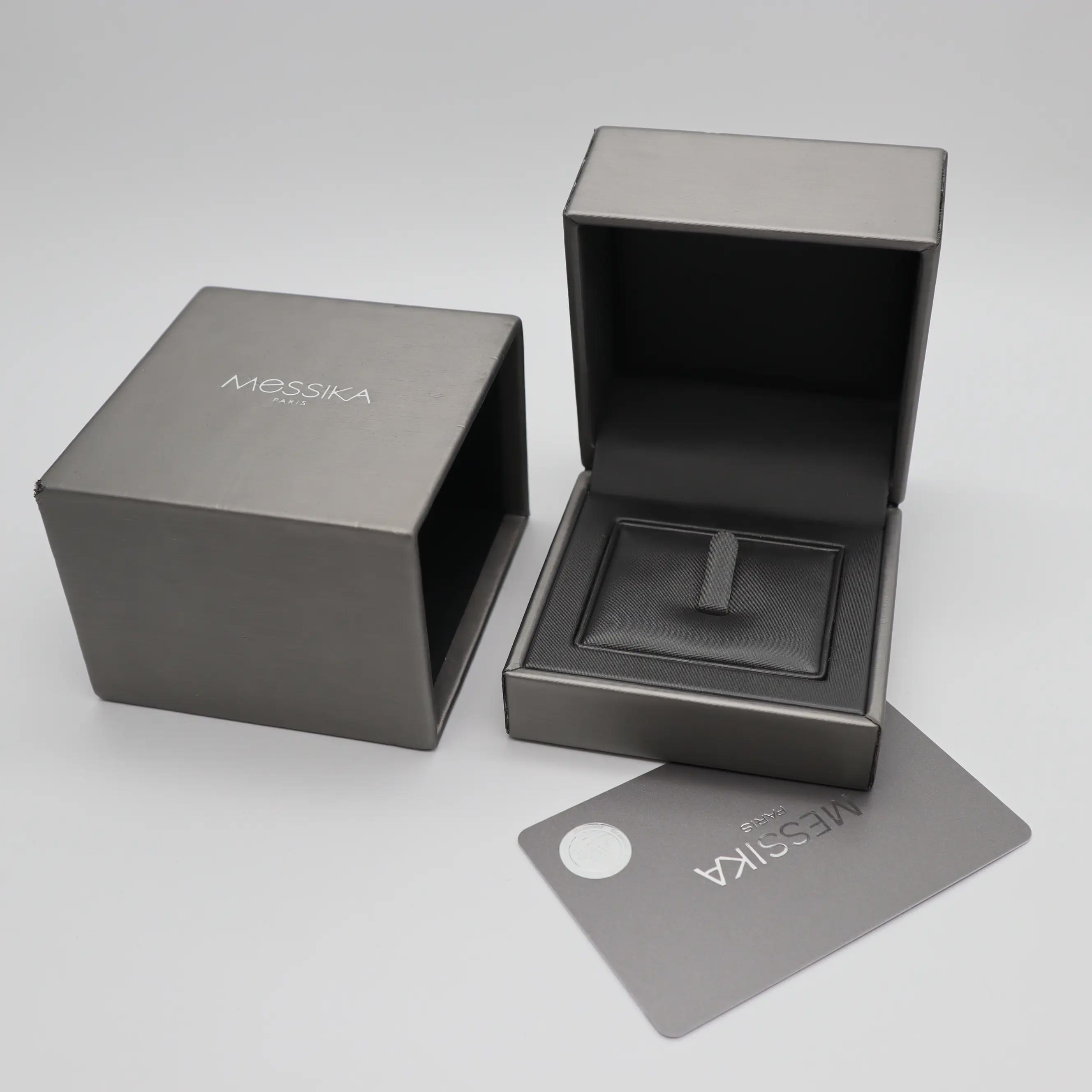 An image of a Messika branded women's ring with diamonds, although the ring itself is not visible. The photo shows a black ring box open and angled at approximately 45 degrees, with the interior and ring holder visible but empty. To the left is a matching closed box, slightly out of focus, and in the foreground, there's a grey Messika card angled away from the camera. The boxes and card are set against a neutral background, and the focus is on the open box, suggesting the product should be there.