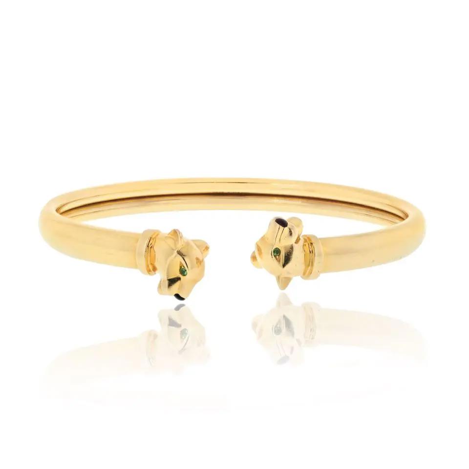 An image of a Cartier unisex adult bracelet with gemstones, showcased against a white background. The bracelet is photographed from a slightly elevated angle, centered in the frame with a soft reflection on the surface below. It features a sleek, polished yellow gold open bangle with a slight oval shape, and is capped on each end with detailed gold panther heads, each embellished with Tsavorite eyes and Onyx nose gemstones. The design is elegant, with the bracelet appearing to float above the reflective surface, positioned horizontally across the image. The distance from the camera provides a clear view of the entire piece, highlighting its luxury and craftsmanship. Great preowned condition.