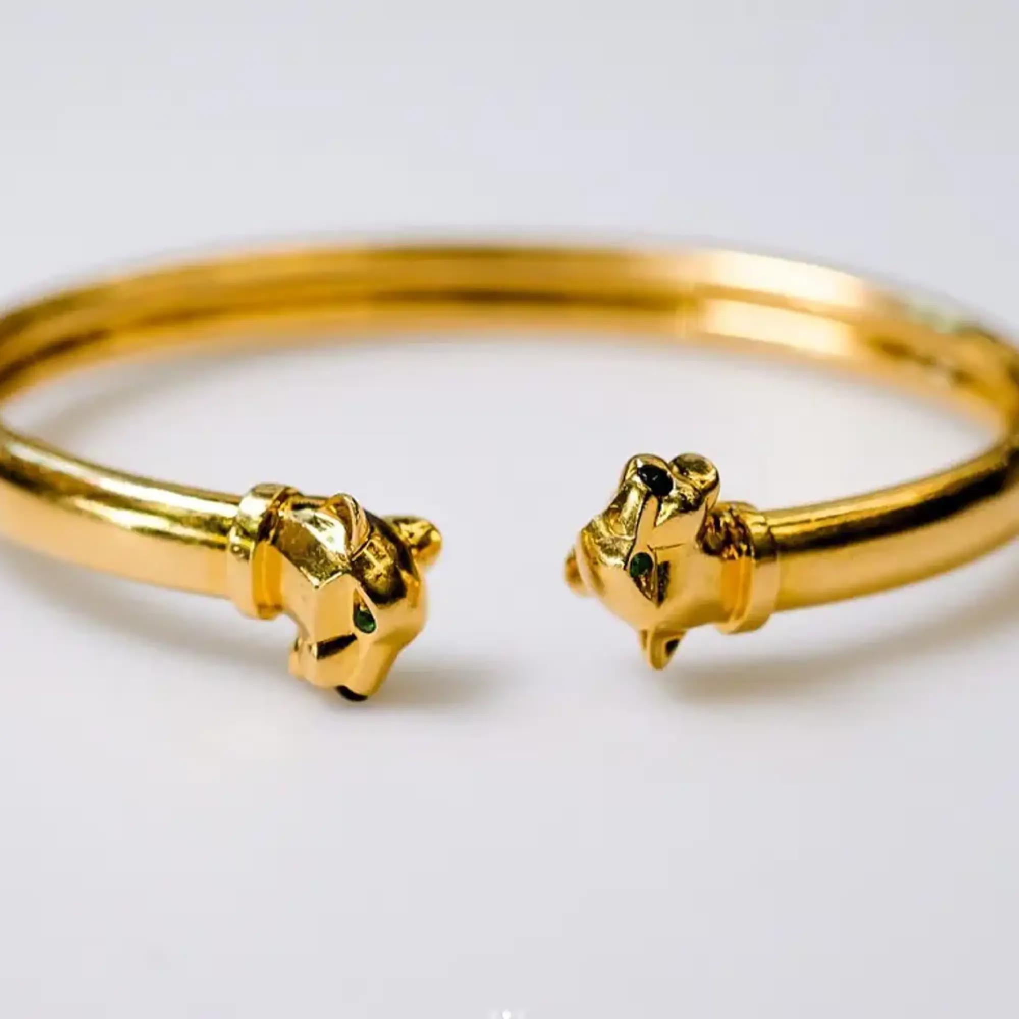 An image of a unisex adult Cartier bracelet with gemstones. The bracelet is positioned horizontally across the center of the frame with a close-up view. It features a polished yellow gold finish with two detailed panther heads facing each other at the ends. Each panther head is accented with small Tsavorite and Onyx gemstones for eyes and nose. The angle of the photo captures the intricate details and the lustrous sheen of the yellow gold metal. Great preowned condition.