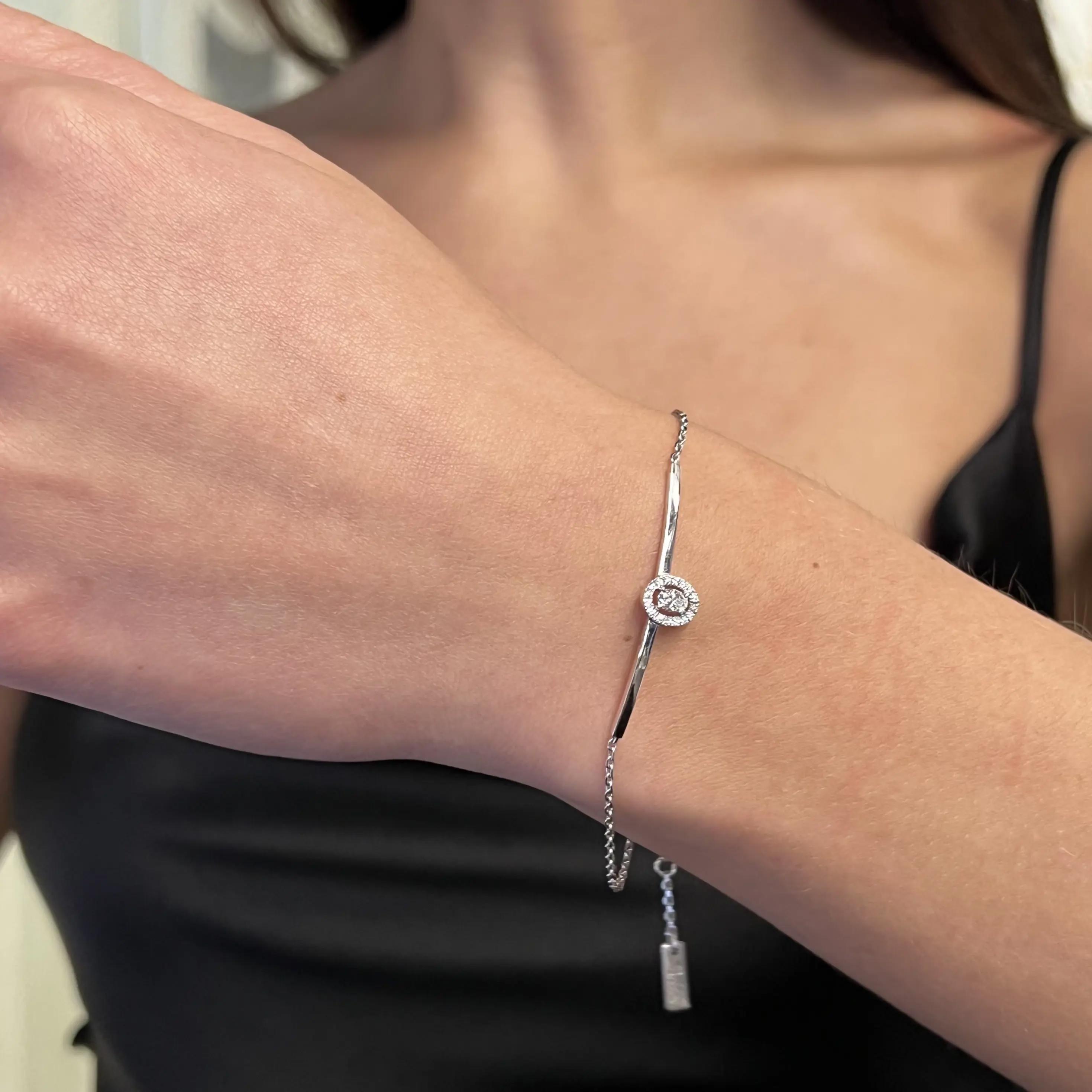 An image of a slender Messika bracelet with diamonds, designed for women, displayed on a wrist. The bracelet is positioned diagonally across the center of the frame, with a close-up view that emphasizes the sparkling central diamond surrounded by smaller diamonds. The bracelet's chain extends towards the bottom right corner of the image, with a small, rectangular Messika brand tag visible near the clasp. The background is out of focus, highlighting the jewelry's details.