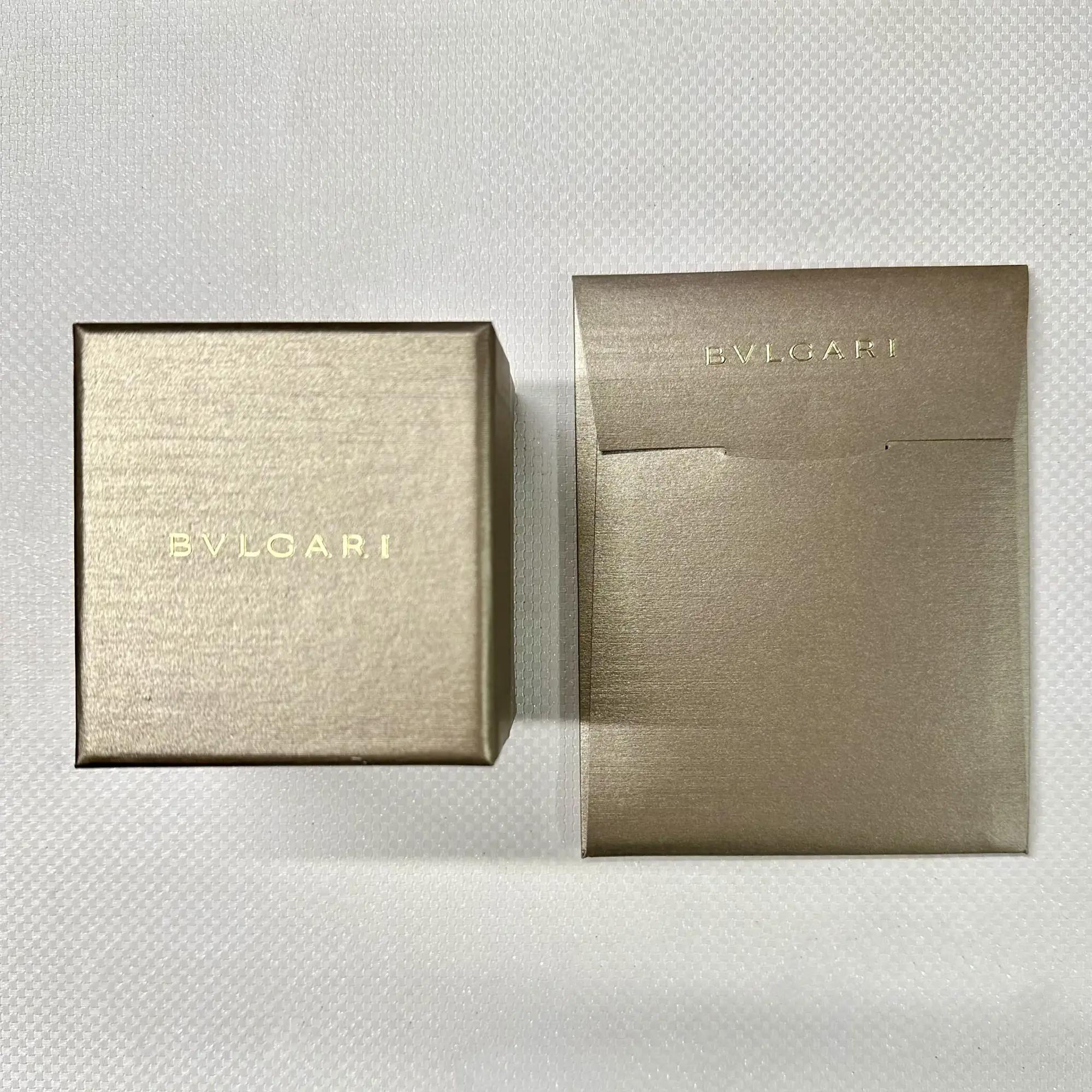 An image of a Bvlgari branded ring box and packaging, presented on a textured surface. The ring box, positioned on the left, is square, with a beige-gold finish and the Bvlgari logo embossed in the center. To the right, there is a rectangular packaging piece, standing upright with a matching beige-gold hue and the Bvlgari logo at the top. The items are captured from a top-down angle, with the camera at a close distance, providing a clear view of the textures and branding. The actual ring with diamonds is not visible in this image.
