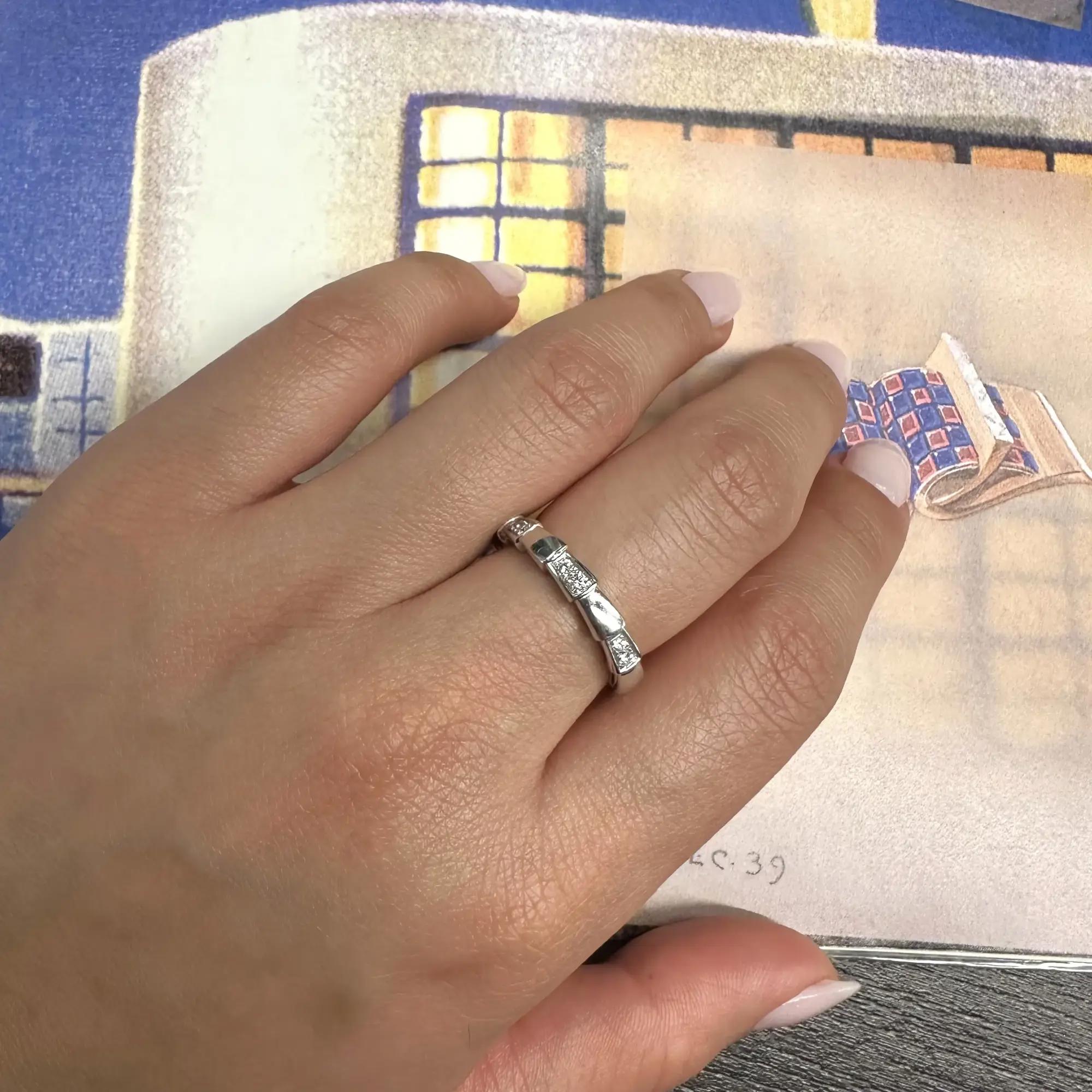 An image of a unisex adult Bvlgari Serpenti Viper band ring with diamonds, worn on the ring finger of a left hand which is resting against a colorful, illustrated background. The hand is positioned at a three-quarter angle from the viewer's perspective, with the diamonds on the ring catching light and sparkling. The ring is shown in a close-up view, allowing for detailed visibility of the diamond settings and the band's design. The condition of the ring is new.