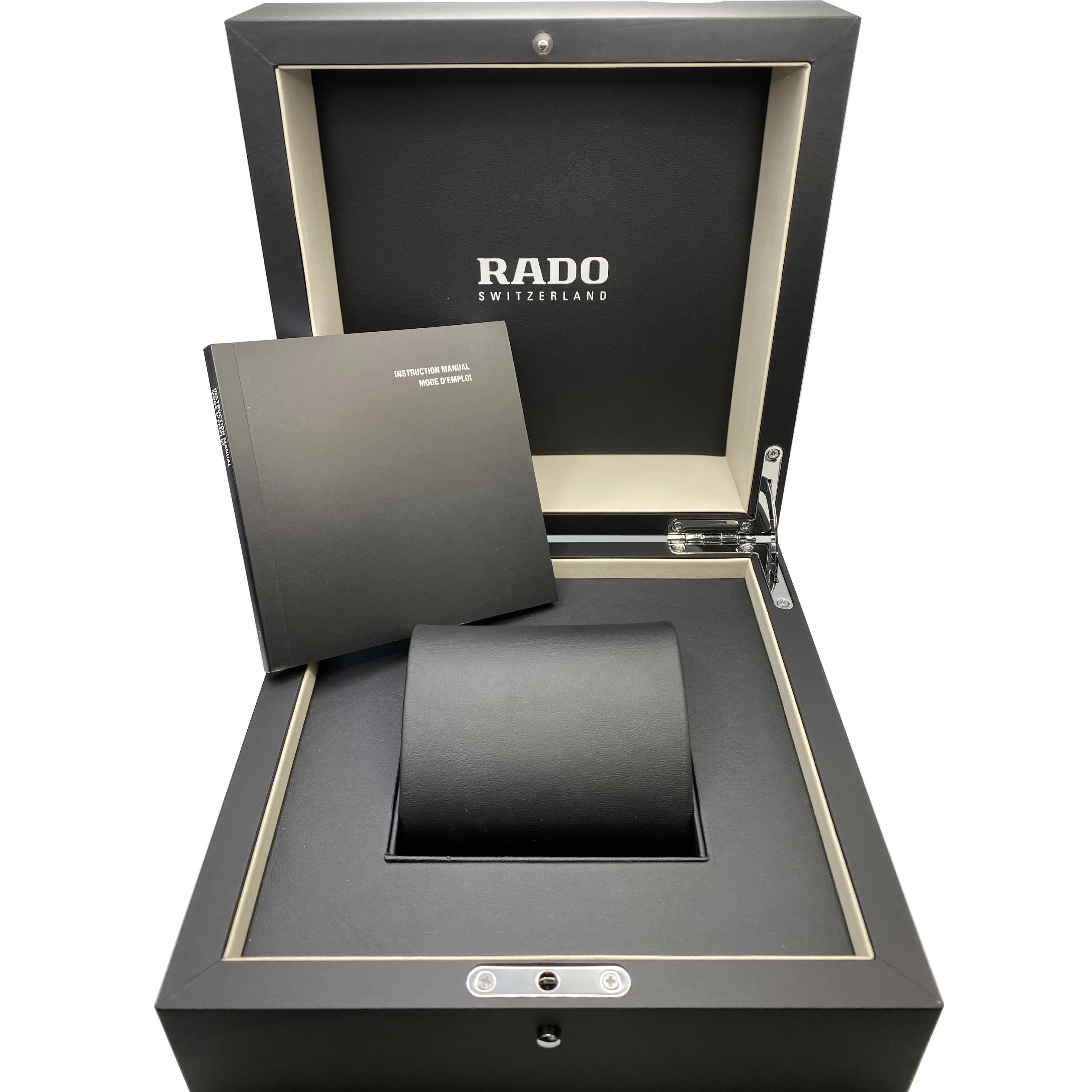 An image of an open Rado wristwatch box for women, viewed from a high angle. The box is black with the Rado logo in silver at the top of the inside lid. Inside the box, positioned centrally, is a cushion for the watch, but the watch itself is not present. To the left, there is a black instruction manual with silver text. The box and documents appear unworn and are in full view, providing a sense of the brand's presentation for a new timepiece. The image is taken from a close distance, focusing on the details inside the box.
