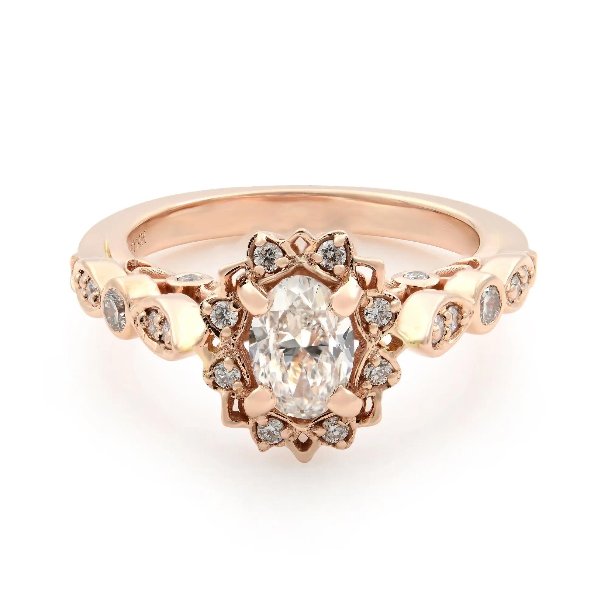An image of a Rachel Koen brand engagement ring for women, showcasing a central oval-cut diamond set in a rose gold band. The ring is positioned at a slight angle to the camera, allowing for a clear view of the intricate setting and the smaller round diamonds encircling the main stone. The image is taken from a close-up perspective to highlight the ring's details, and the lighting reflects off the diamonds to emphasize their brilliance.
