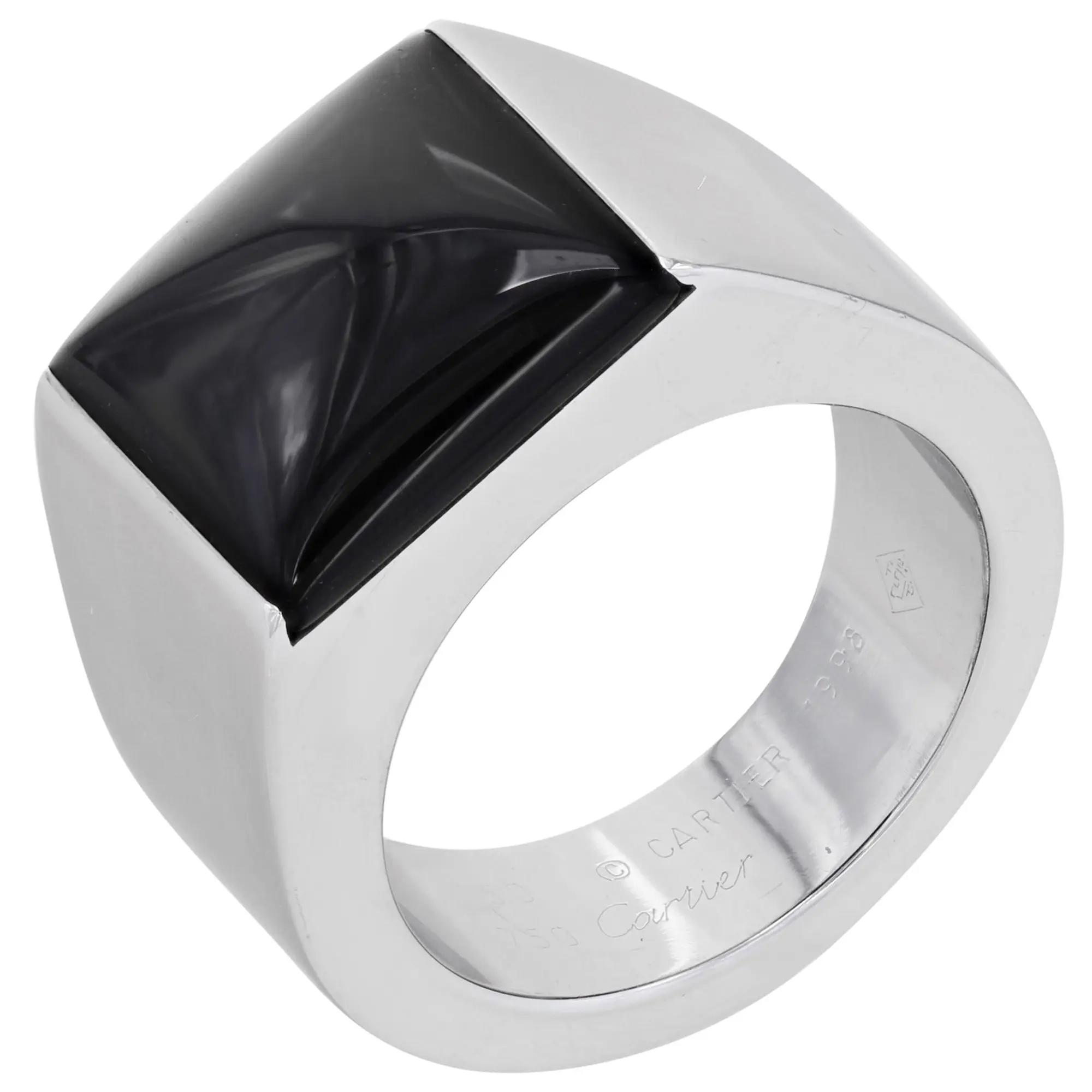 An image of a unisex adult Cartier ring with gemstone, positioned at a slight angle to showcase the top and side view. The ring features a prominent black Onyx gemstone set in a thick white gold band. The gemstone has a pyramidal cut and is mounted flush with the band's surface. The Cartier brand name is engraved inside the band, visible on the lower right side of the image. The photo is taken from a close distance, ensuring that the details of the gemstone and the engravings are clear. Great preowned condition.