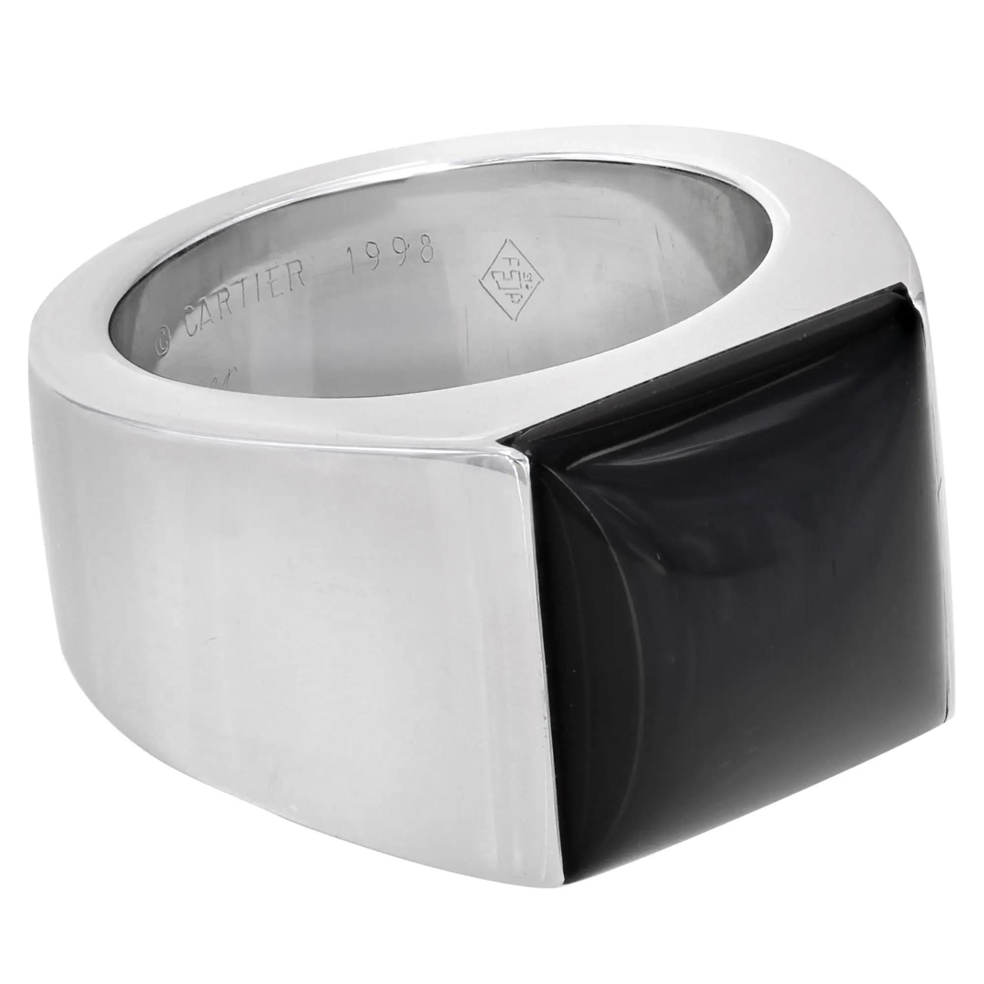 An image of a Cartier unisex adult ring with gemstone, shown in a close-up side view. The ring features a bold, square design with a polished platinum band and a large black Onyx gemstone occupying half of the front face. The Cartier logo and the year "1998" are engraved on the inside of the band, visible from the angle. The gemstone has a smooth, glossy finish and contrasts strikingly with the white gold metal. The image is taken from a slightly elevated angle, focusing on the gemstone and the engravings inside the band. Great preowned condition.