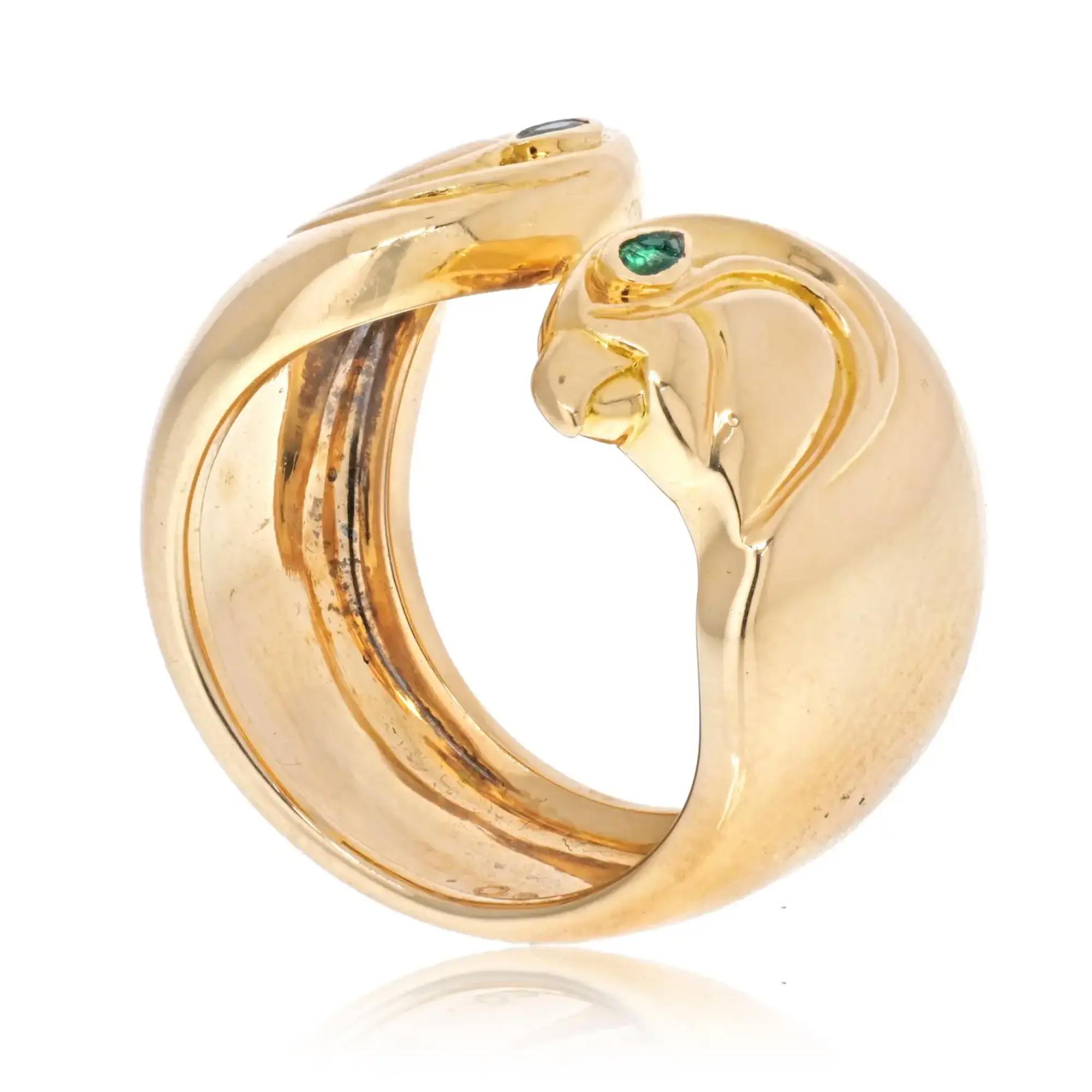 An image of a women's Vintage Cartier Anoubois ring with gemstones, positioned upright and viewed at a slight angle to showcase the design and gemstone placement. The ring is in yellow gold, featuring a thick band that splits into two curved sections at the top. A small, round green gemstone is embedded in one section, close to where the split begins. The light reflects off the metallic surface, highlighting the ring's polished finish and the subtle wear marks indicating usage. The shot is taken at a close distance, focusing on the ring to capture its details against a white background. Comes in a preowned condition.